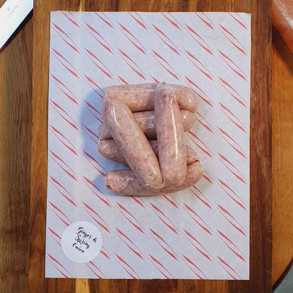 6 chunky handmade pork, ginger & spring onion sausages on waxpaper surrounded by Himalayan salt blocks and butchers tools on a wooden bench.